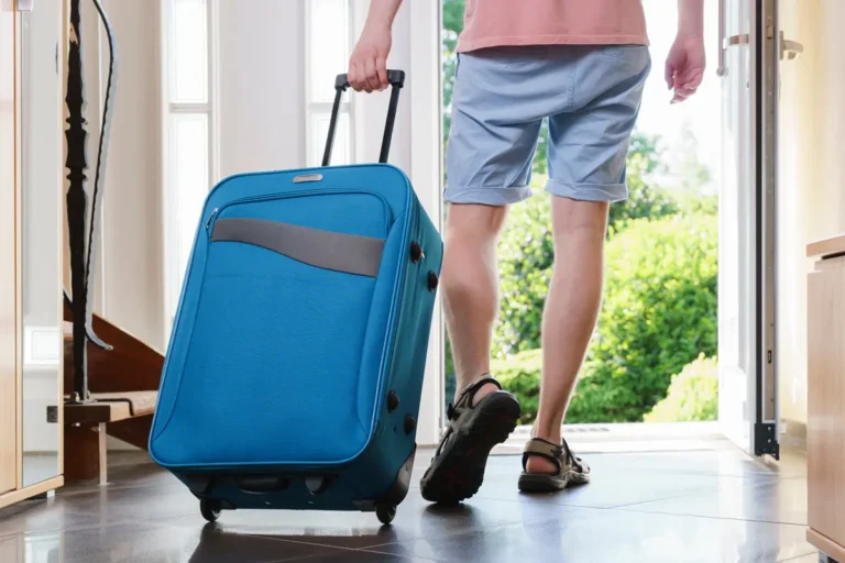 Person leaving home with a suitcase, walking toward the front door on a bright summer day.