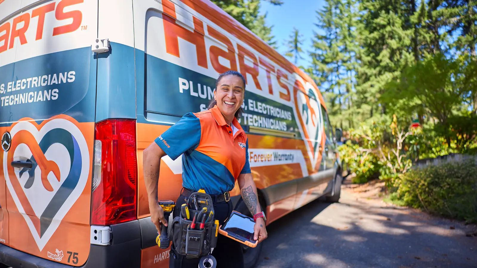 Harts technician standing with tools and a tablet in front of a branded Harts Plumbers, Electricians, & HVAC Technicians van.