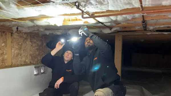 Plumbers inspecting pipes under a home during an emergency plumbing repair call.
